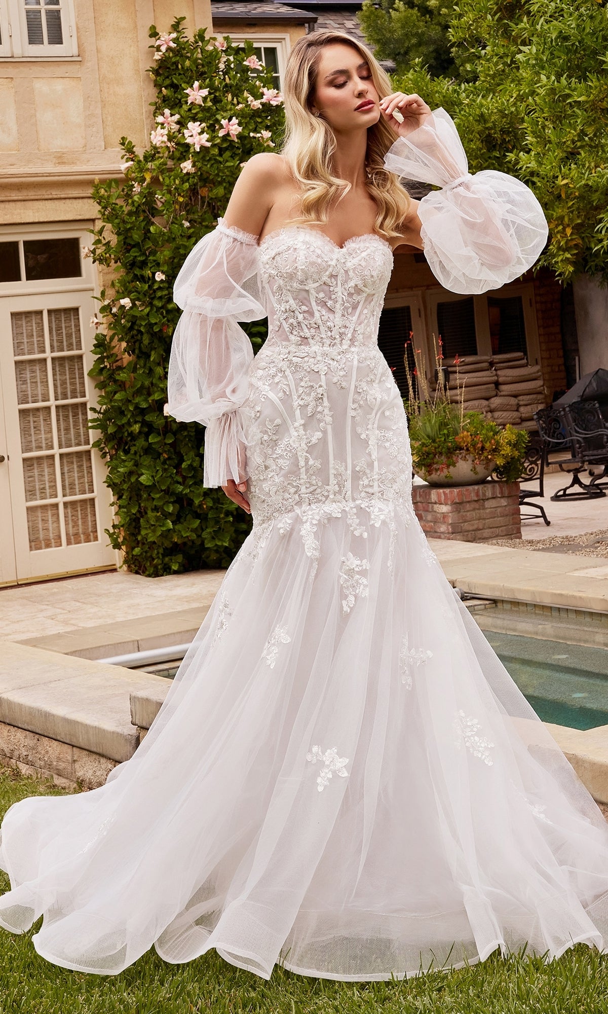 A woman stands outdoors wearing the Puff Sleeve Mermaid Wedding Gown CD858W, featuring white lace, tulle, floral appliqués, and removable puff sleeves.
